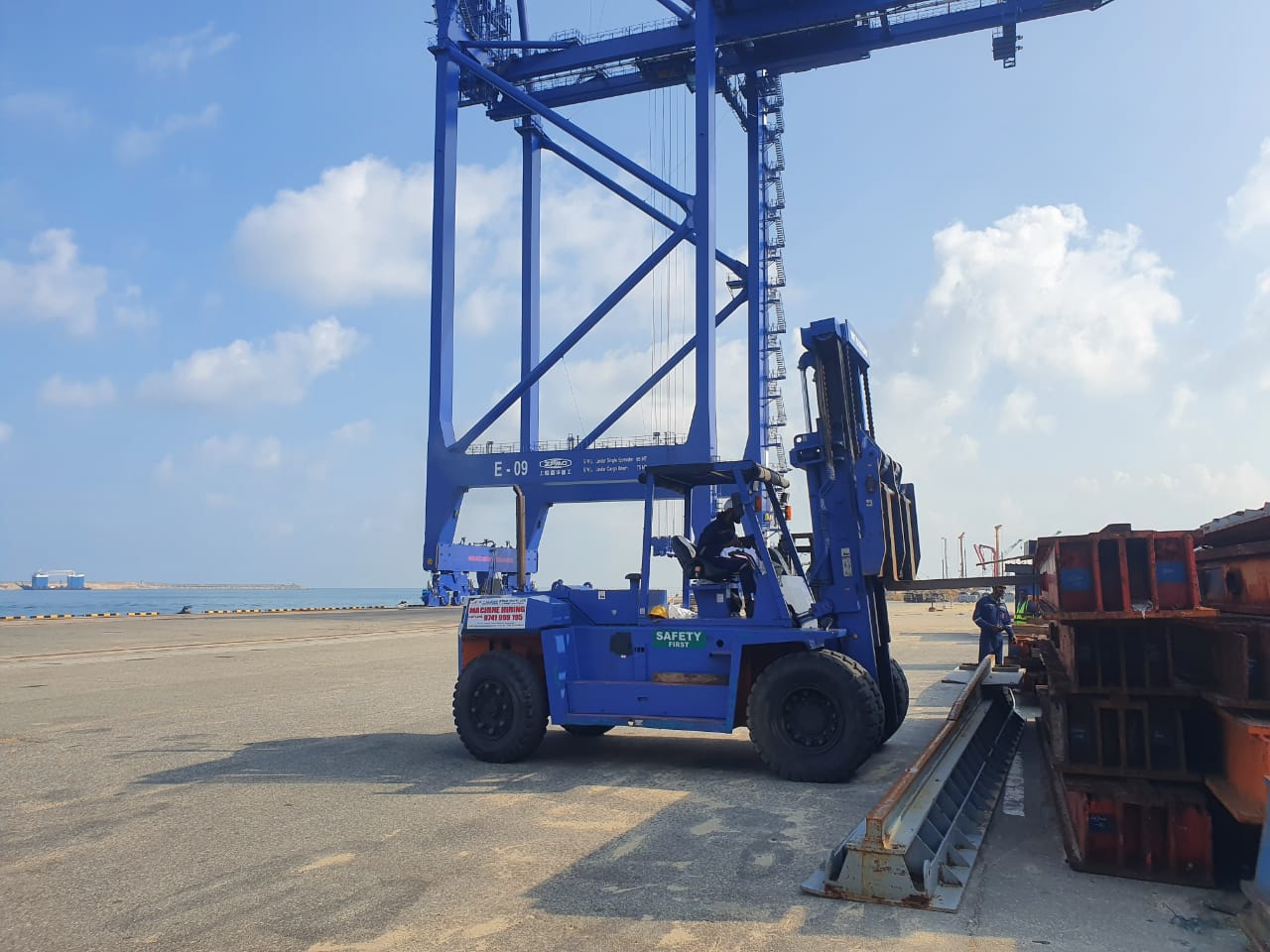 Blue forklift operating near stacked metal containers and a large crane on a dock under a blue sky.