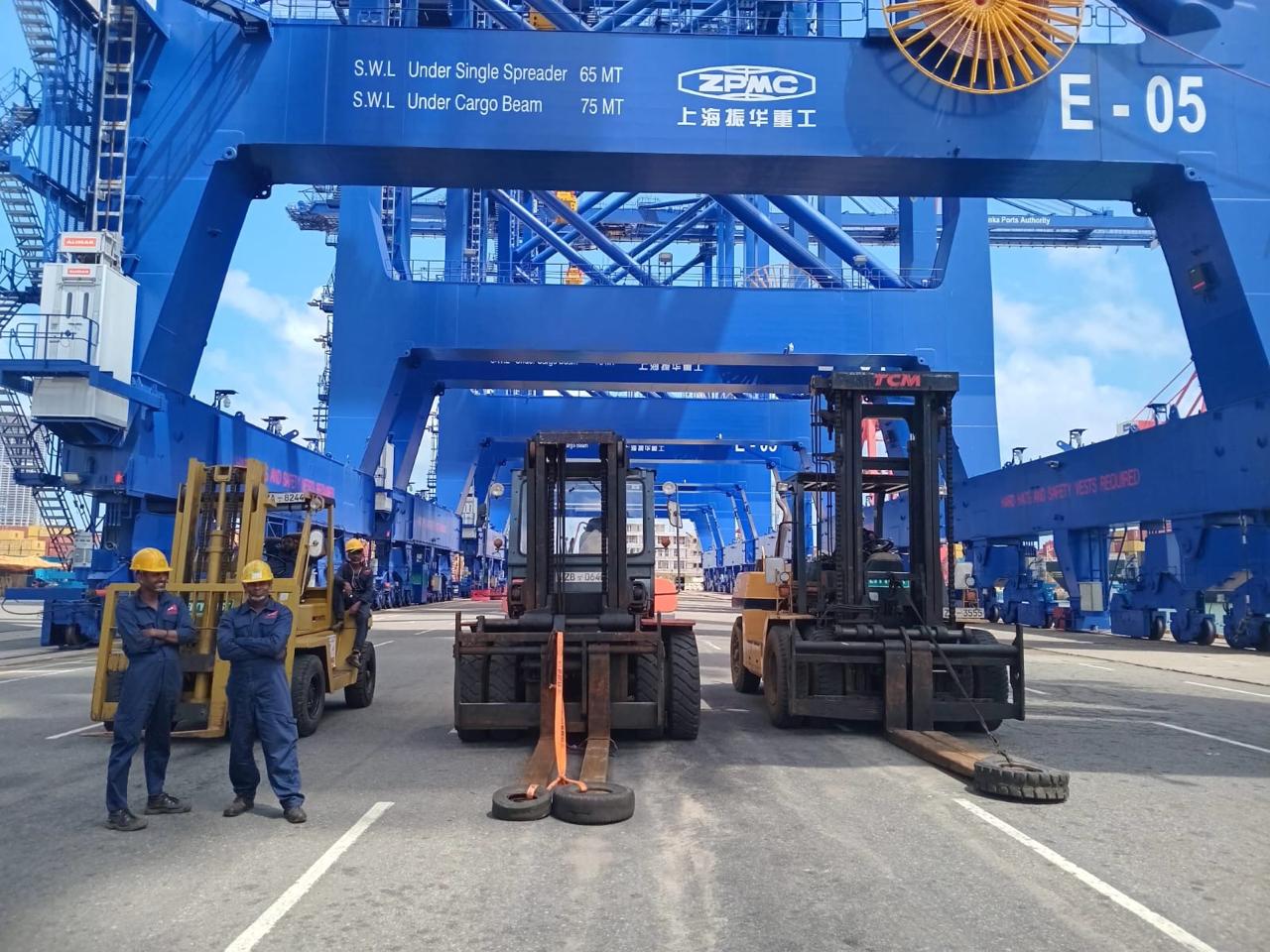 Three forklifts parked under large blue cranes with two workers in yellow helmets standing nearby.