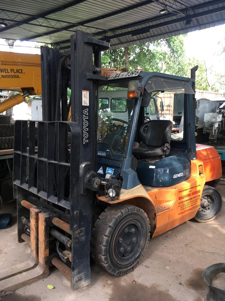 Orange and black Toyota Geneo 35 forklift parked under a metal roof with trees in the background.