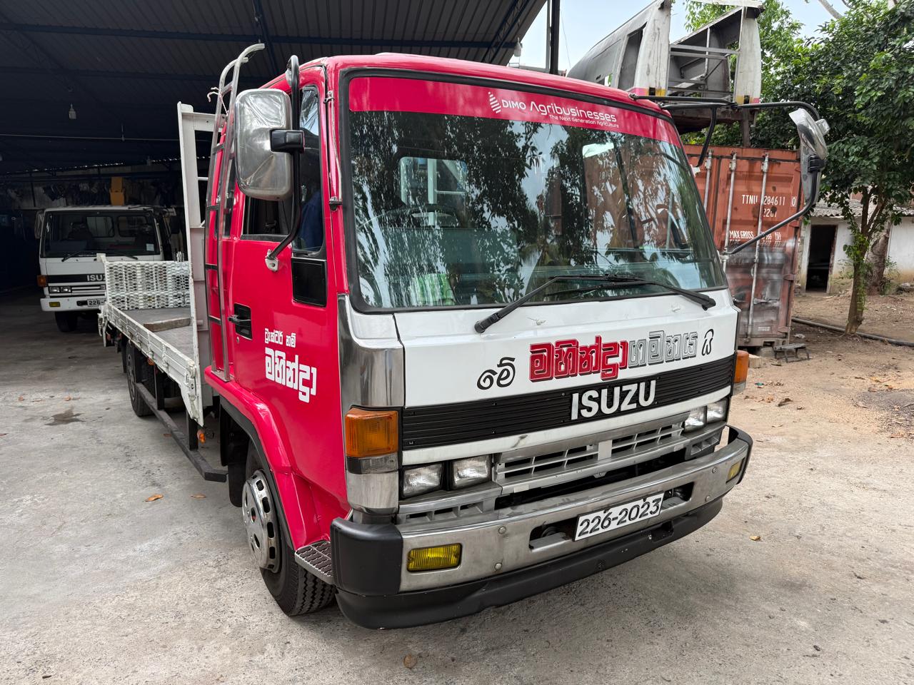Red and white Isuzu flatbed truck with advertising text parked in a garage with another truck behind.