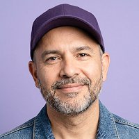 Smiling middle-aged man with a beard wearing a purple cap and denim jacket, purple background.