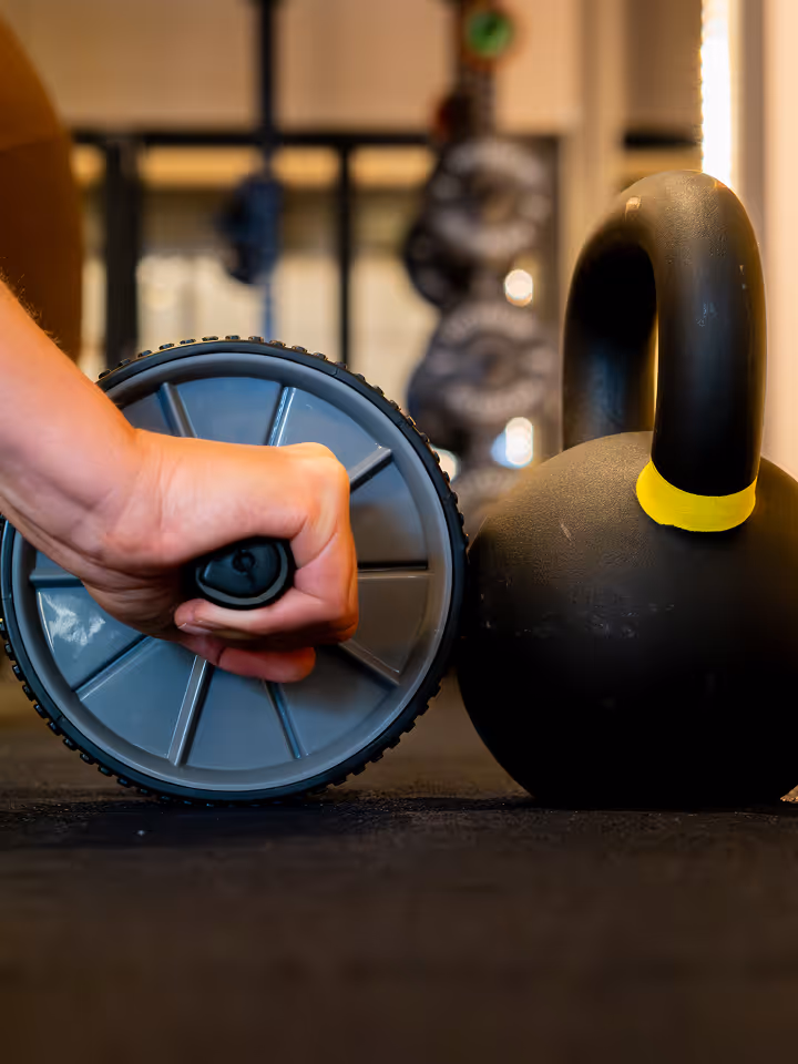Hand die een grijze fitnesswiel vasthoudt naast een zwart kettlebell met een gele band op een sportvloer.