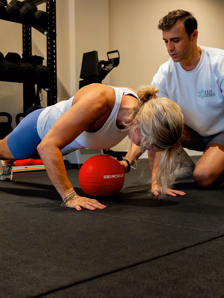 Vrouw die push-ups doet met hand op een rode Rogue-medicijnbal terwijl een trainer toekijkt in een sportschool.