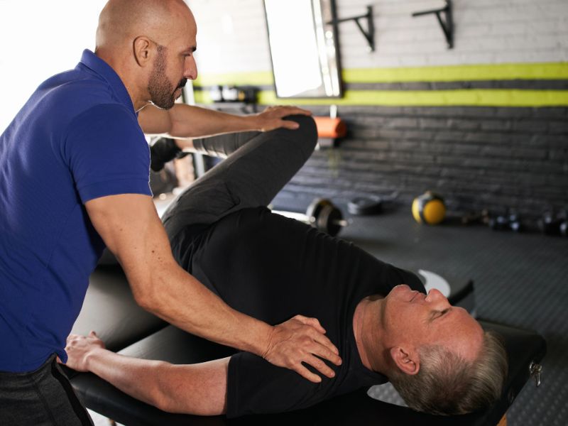 Therapist assists older man lying on table with leg stretch in a gym setting.