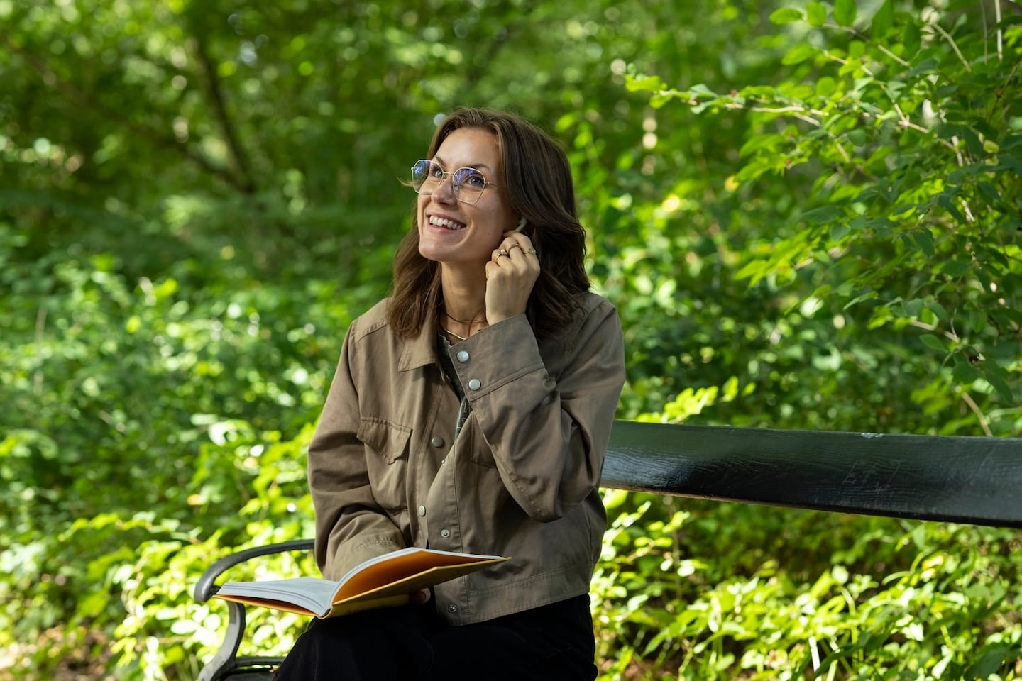 Vrouw met een bril die op een bank in een groen park zit en glimlacht terwijl ze naar boven kijkt en een boek vasthoudt.
