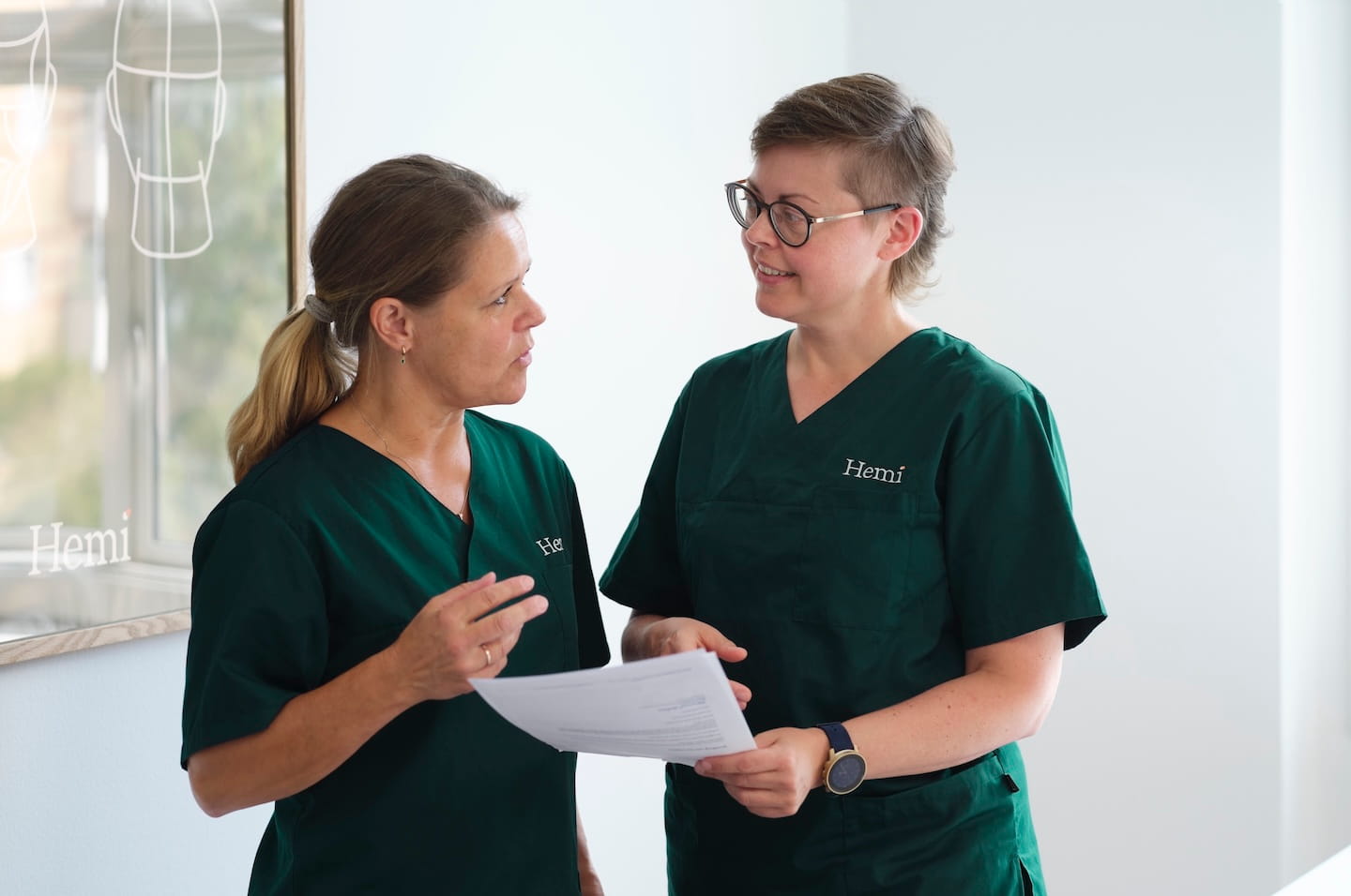 Twee medisch personeel in groene uniformen van Hemi bespreken documenten in een lichte ruimte.