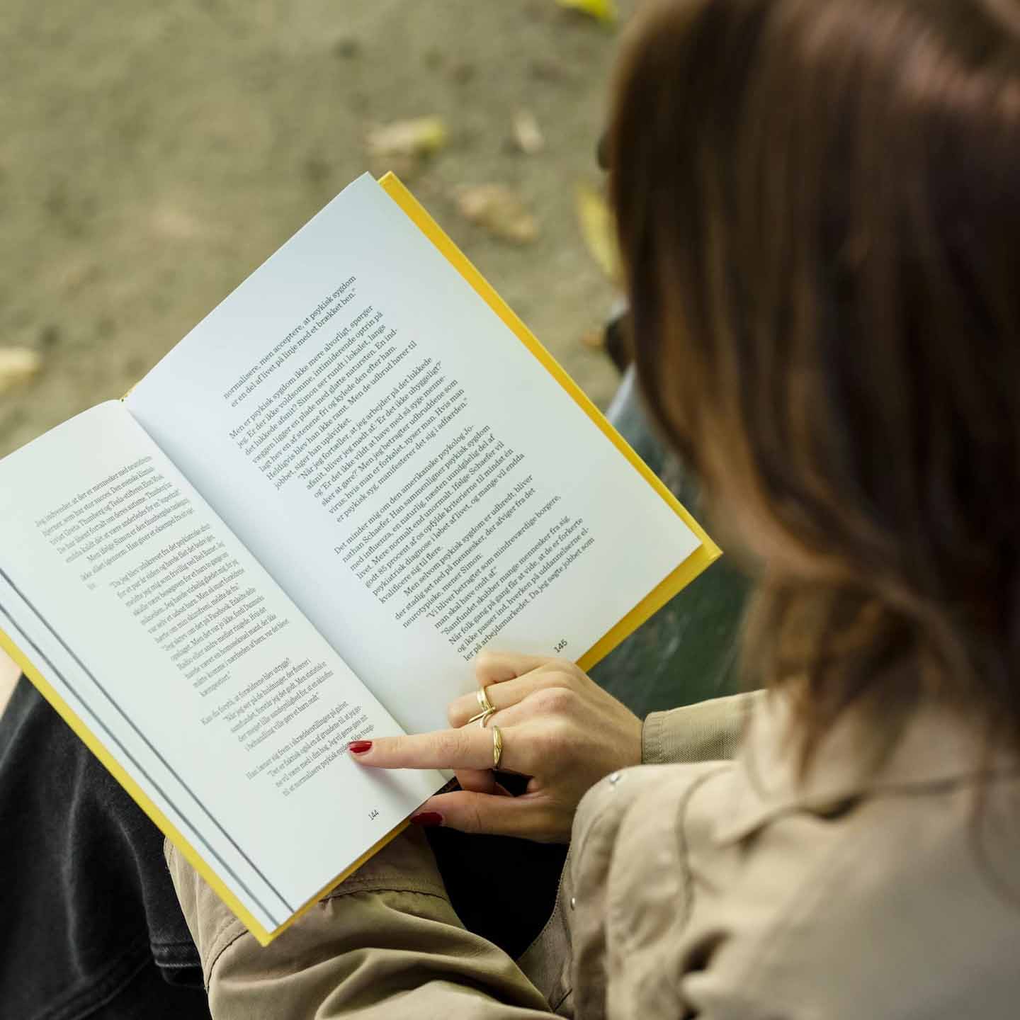 Persoon met beige jas die een open boek leest in een buitenomgeving, wijzend op een specifieke regel tekst.