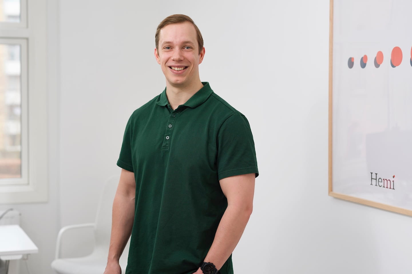 Man in een groen poloshirt glimlacht staand in een lichte kamer met een raam en een wit bord aan de muur.
