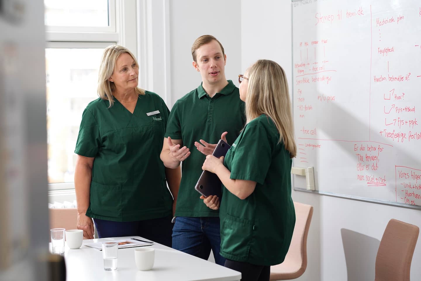 Drie mensen in groene uniformen overleggen voor een whiteboard met aantekeningen in een vergaderruimte.