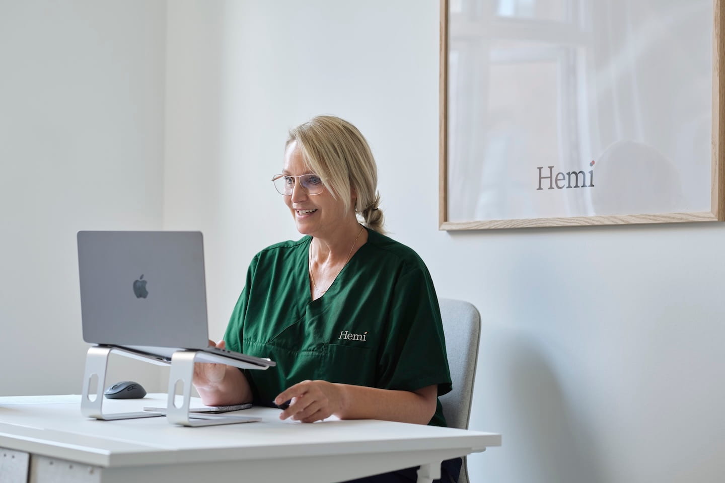 Vrouw in een groen uniform met 'Hemi' op de borst werkt op een laptop aan een witte tafel in een lichte kantoorruimte.