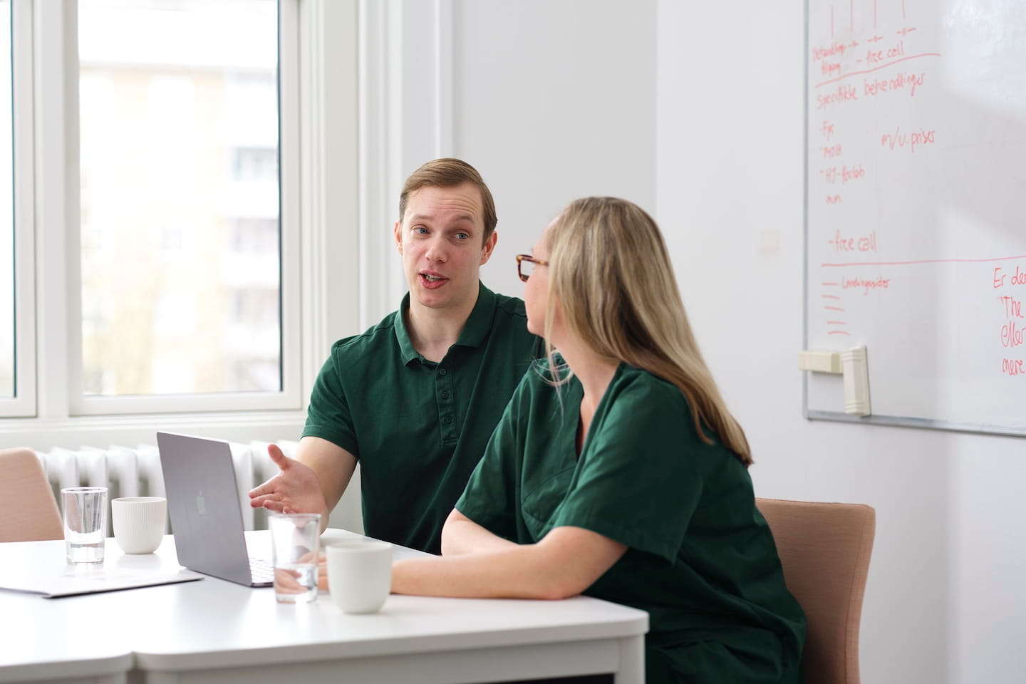 Twee mensen in groene werkkleding zitten aan een tafel met een laptop, met een whiteboard op de achtergrond.