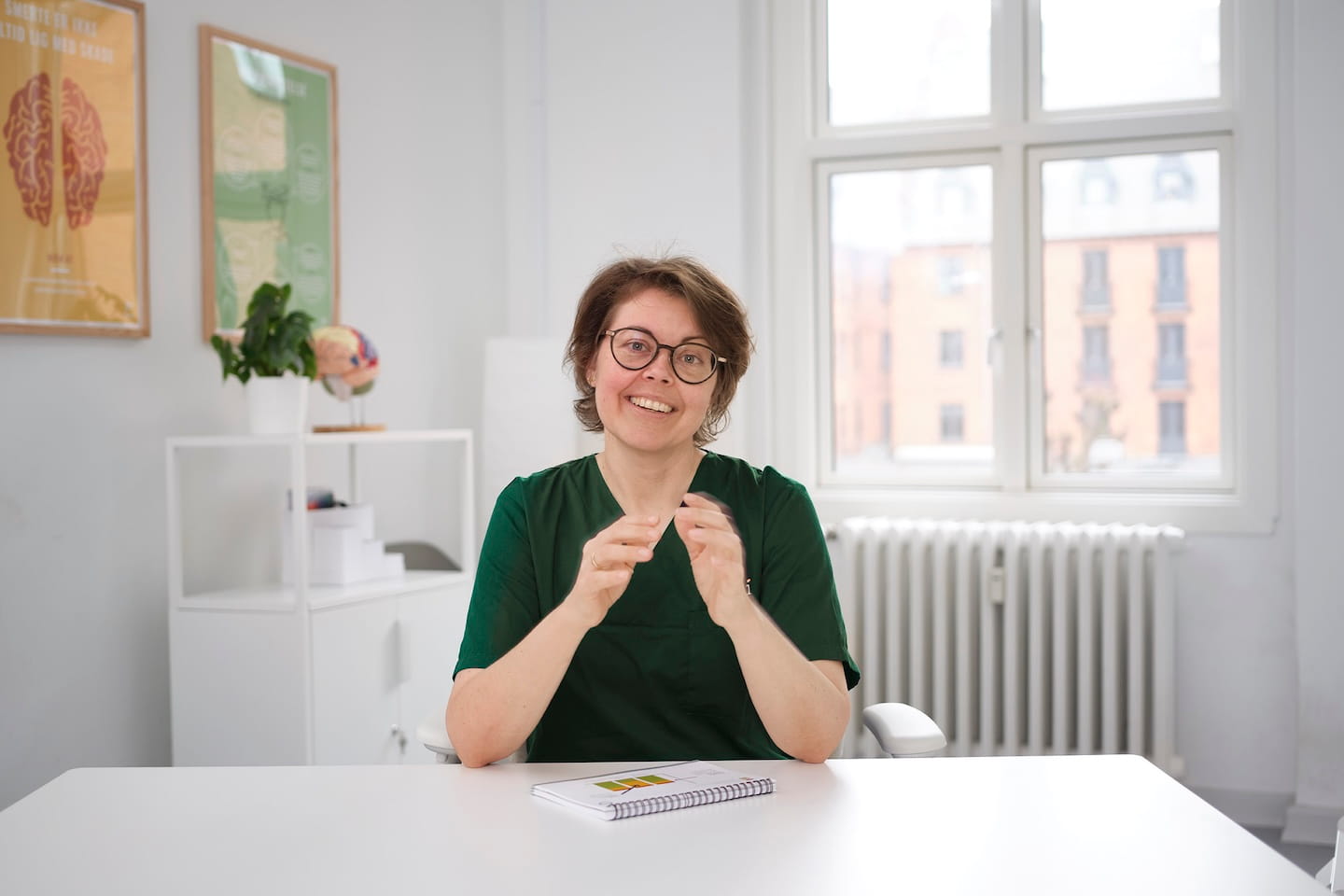 Een lachende vrouw met een groene artsenjas en bril zit aan een wit bureau met een notitieboekje in een lichte kamer met posters aan de muur en een raam.