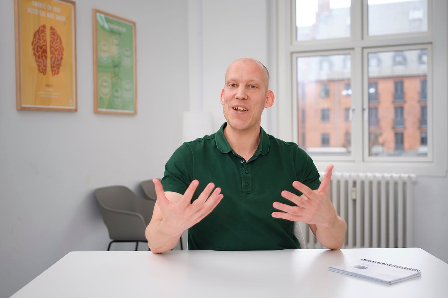 Man in groen poloshirt zit aan tafel en praat met handen terwijl hij in een lichte kamer met raam en posters zit.