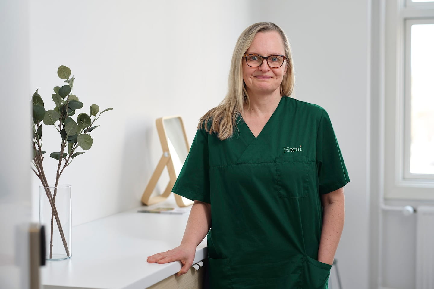 Vrouw met bril en groen uniform met tekst 'Hemi', staand in een lichte kamer bij een wit aanrecht met een glazen vaas met groene takken.