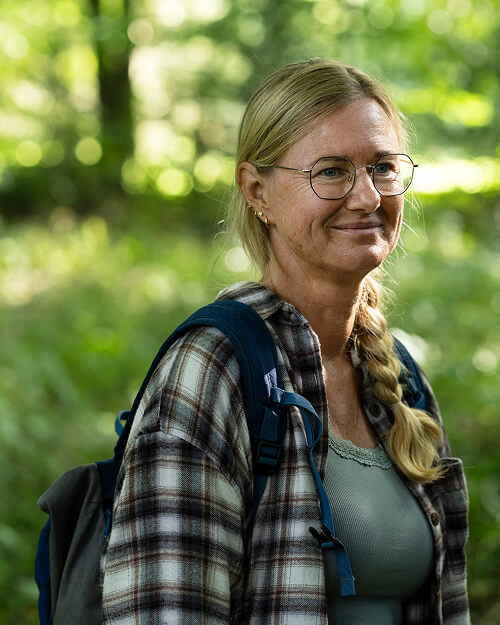 Vrouw met bril en een gevlochten staart, met een rugzak en een geruite jas, die glimlacht in een bosrijke omgeving.