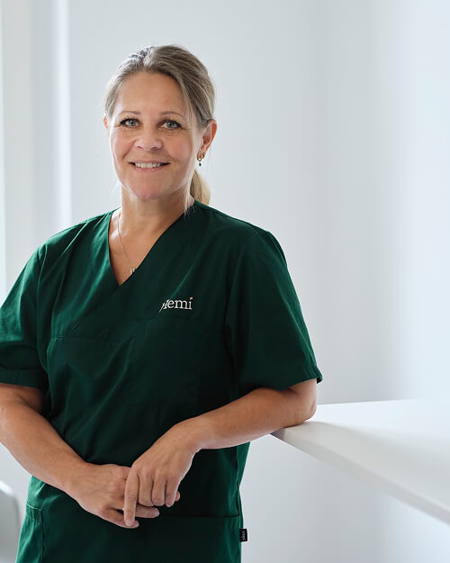 Vrouwelijke zorgprofessional in groen uniform met logo 'jemi', glimlachend en leunend op een witte balie in een lichte ruimte.