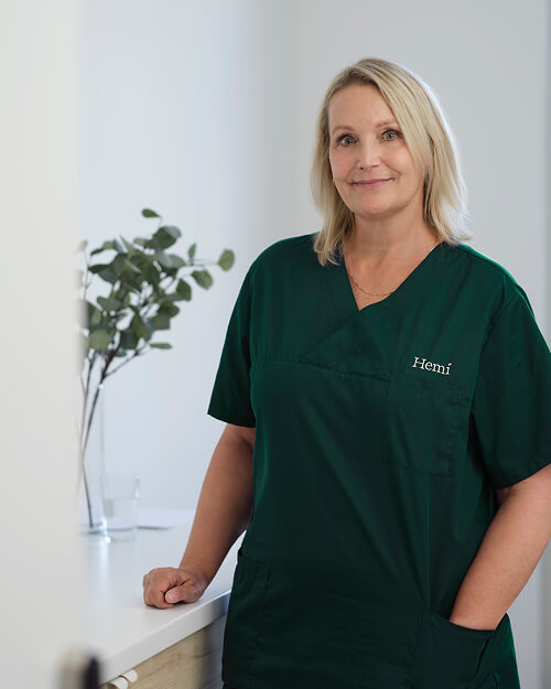 Vrouwelijke zorgprofessional in groene scrub met naam 'Hemi', leunend op een aanrecht met een vaas met groene takken op de achtergrond.