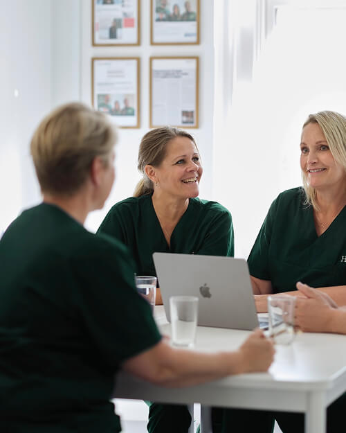 Drie medisch personeel in groene uniformen overleggen rond een tafel met een laptop in een lichte ruimte.