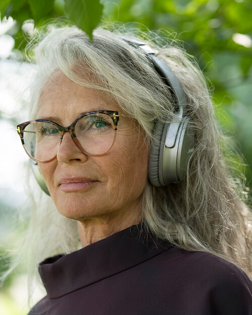Oudere vrouw met grijs haar, bril en grote koptelefoon, buiten tussen groene bladeren.