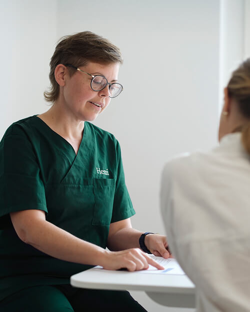 Medisch professional met bril in groene uniform wijst op documenten tijdens consult met patiënt.
