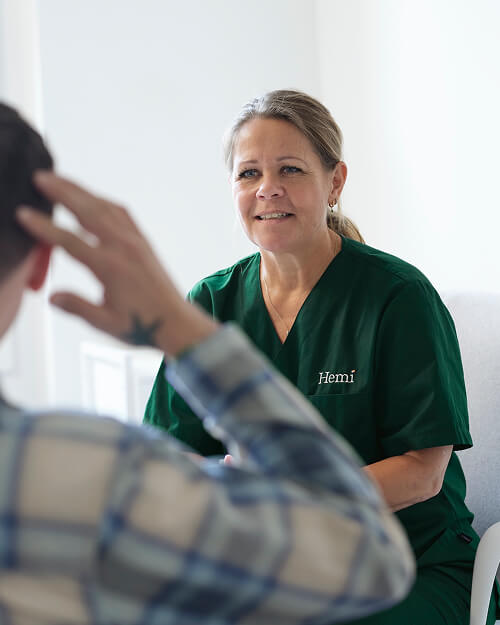 Kvinde i grøn medicinsk uniform med teksten 'Hemi' taler venligt med en mand, der har hånden på hovedet og iført skjorte med tern.