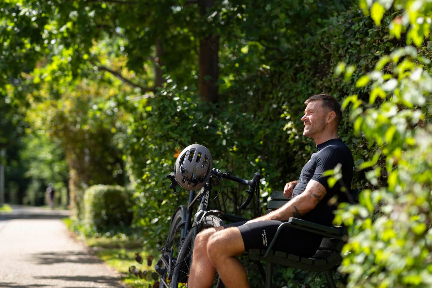 Man in fietskleding zit op een bankje in een groen park naast zijn racefiets met helm.