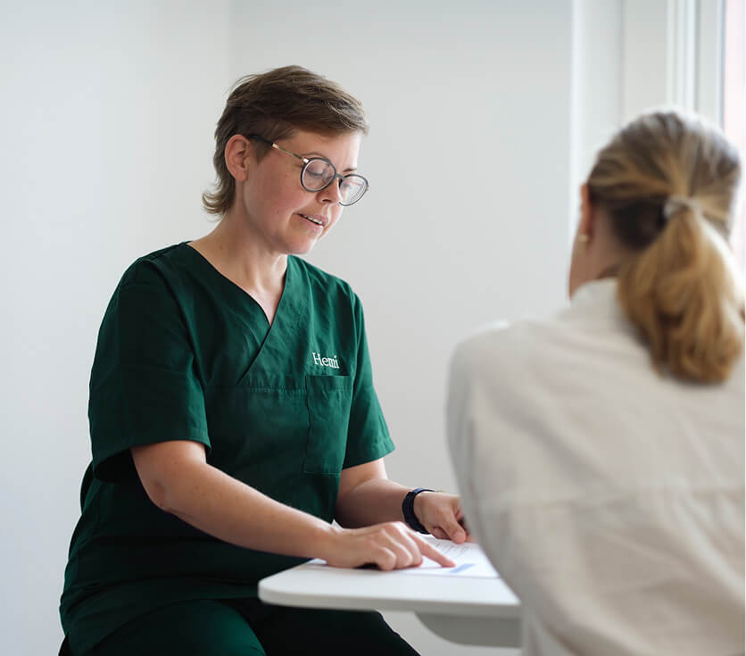 Gezondheidswerker in groene kleding legt informatie uit aan een zittende patiënt in witte kleding.