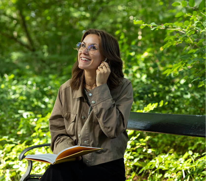 Vrouw met bril zittend op een bankje in een groen park leest een boek en glimlacht terwijl ze haar oor aanraakt.