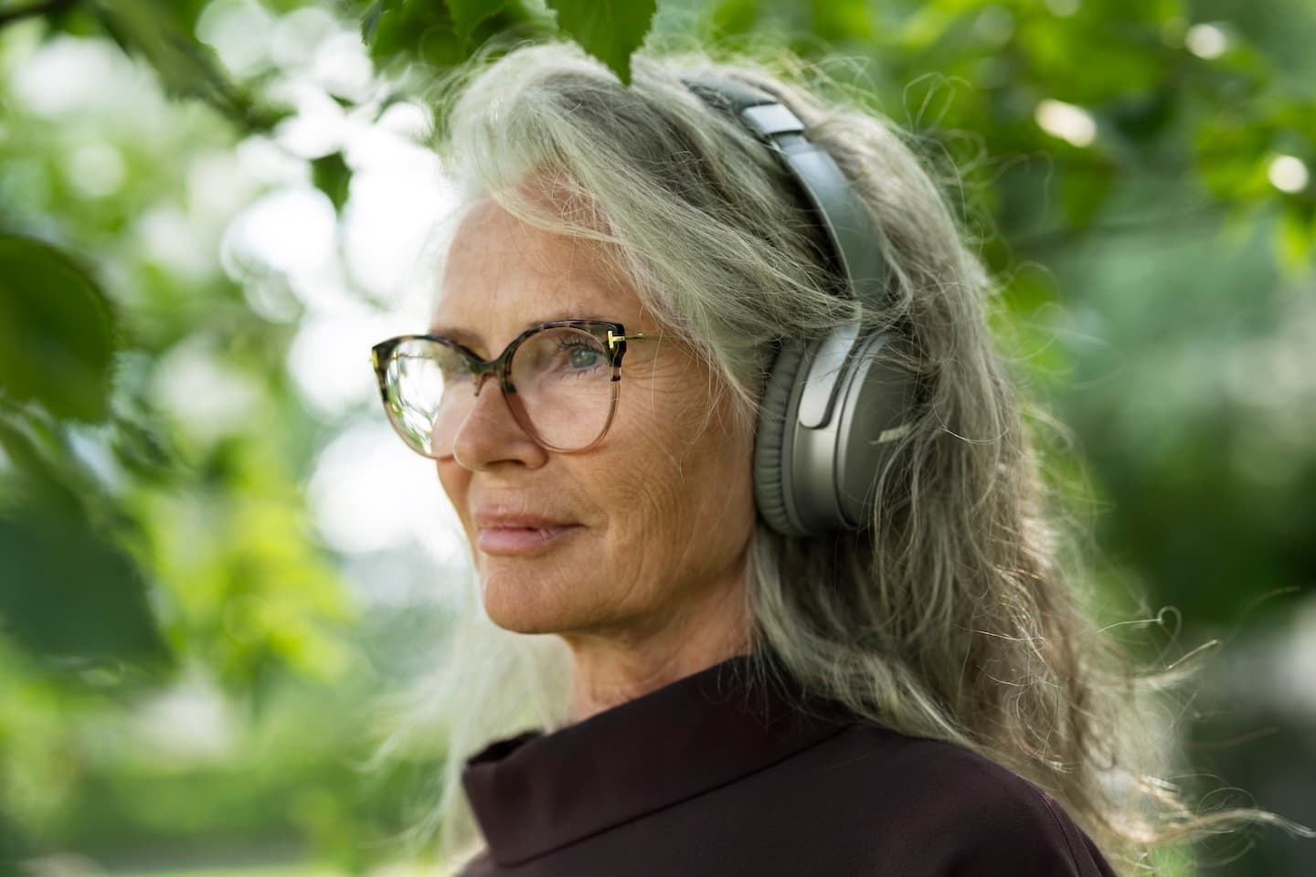 Oudere vrouw met grijs haar draagt een bril en grote koptelefoon, staand in een groene buitenomgeving.