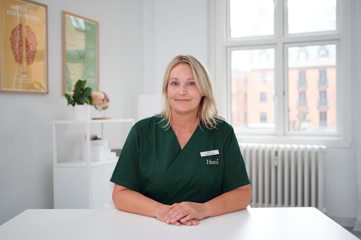 Smilende kvindelig sundhedsarbejder iført grøn uniform med navneskilt, siddende ved hvidt bord i lyst kontor med vindue, radiator og plakater om hjernen i baggrunden.