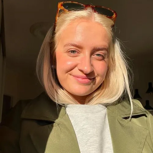 Ung kvinde med lyst hår iført grøn jakke og lysegrå trøje smilende i sollys med briller i håret.