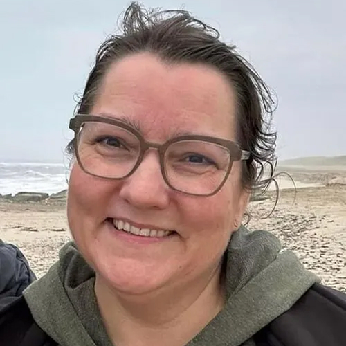 Glad kvinde med briller iført en grøn hættetrøje og sort jakke, smilende foran en strand med hav og sand i baggrunden.