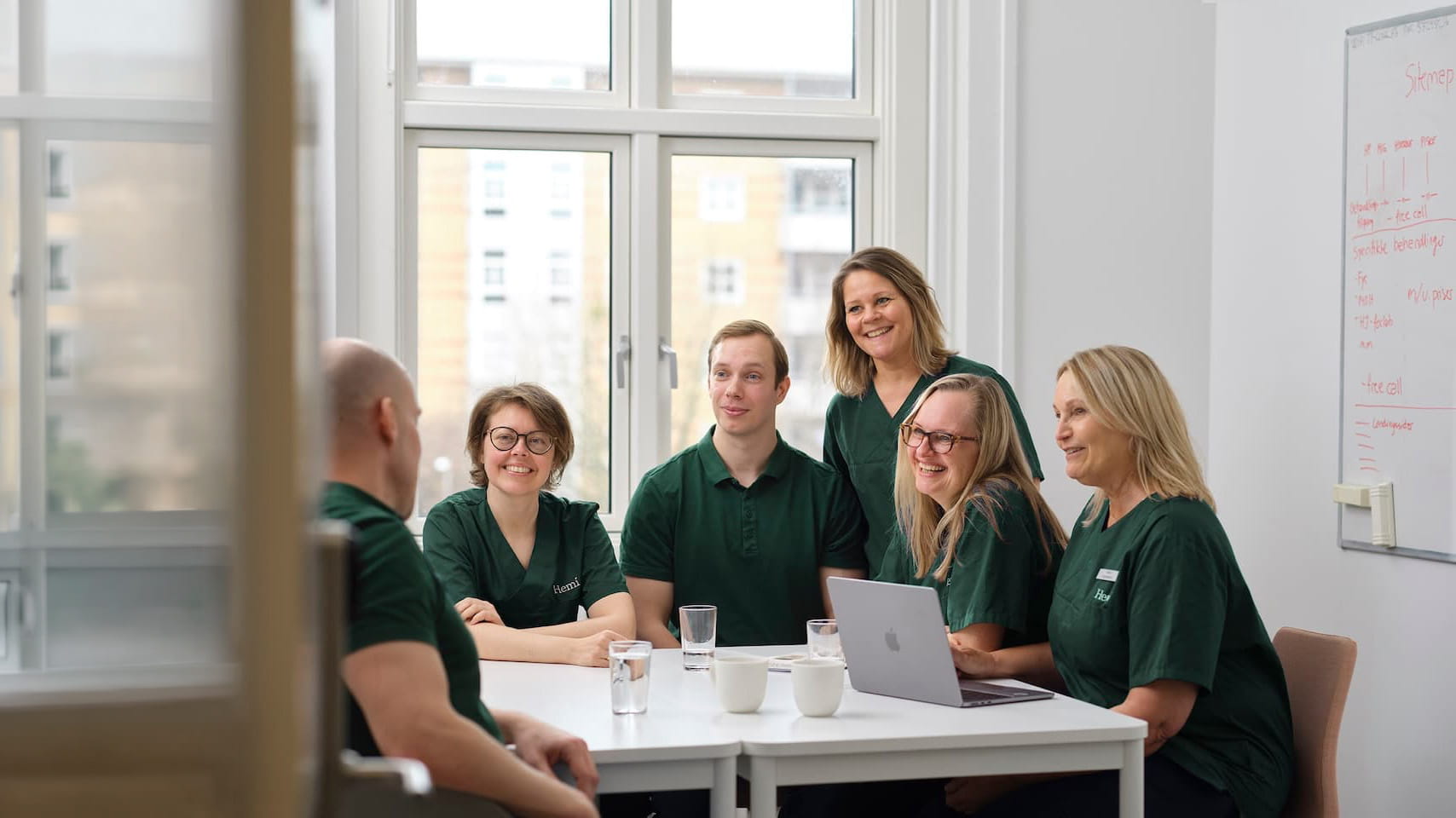 Fem personer iført grønne uniformer sidder rundt om et hvidt bord i et lyst rum med et vindue bag dem og en whiteboard på væggen, de smiler og ser mod en mand med bar hoved i forgrunden.