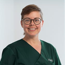 Vrouw met kort bruin haar en bril, glimlachend, gekleed in een donkergroene medische uniform.