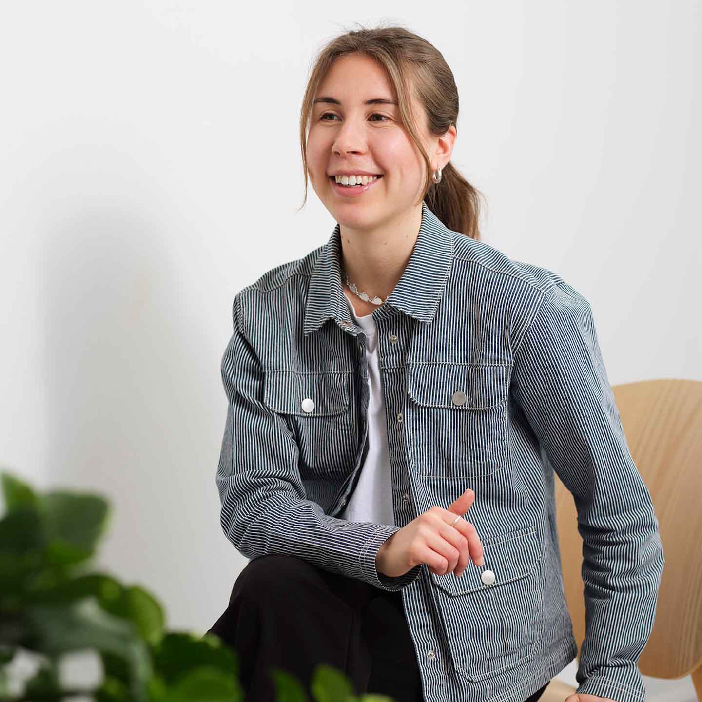 Jonge vrouw met een gestreept jasje, zittend op een stoel en glimlachend, met een groene plant op de voorgrond.