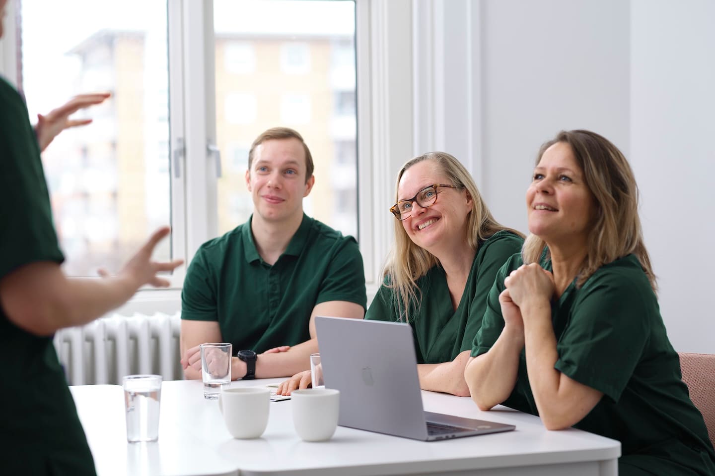 Drie mensen in groene kleding luisteren aandachtig aan een tafel met een laptop en waterglazen tijdens een gesprek.