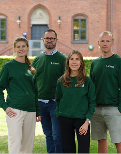 Fire personer iført grønne sweatshirts med 'Hemi' på brystet står foran en murstensbygning med buede vinduer og en sort hoveddør med glasparti.