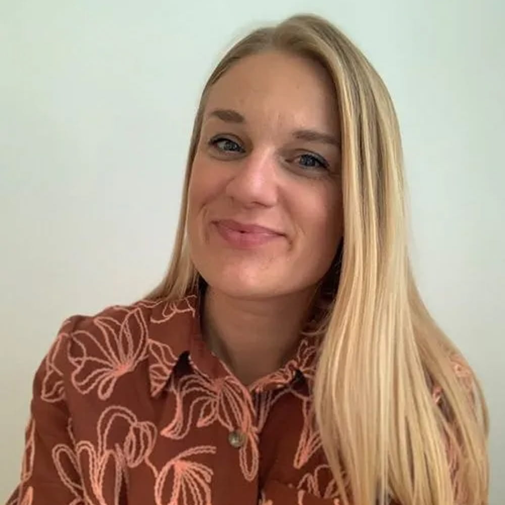 Portræt af smilende kvinde med langt blond hår iført brun skjorte med lyserødt blomsterprint foran en lys baggrund.