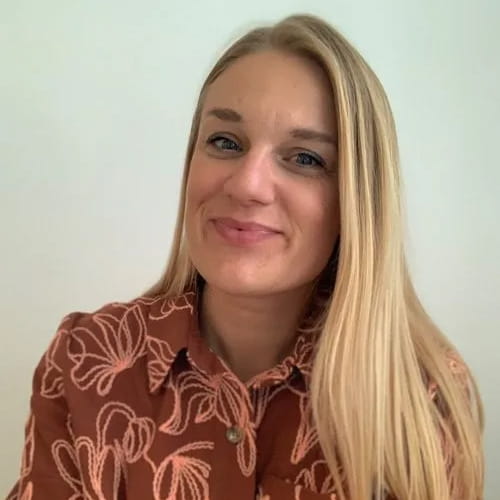 Vrouw met lang blond haar en een bruine blouse met lichtroze bloemenprint, glimlachend naar de camera.