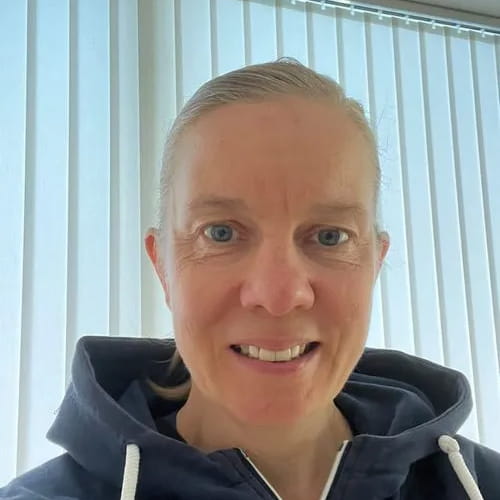 Vrouw met blond haar in een paardenstaart en een donkerblauwe hoodie, glimlachend voor een raam met witte verticale jaloezieën.