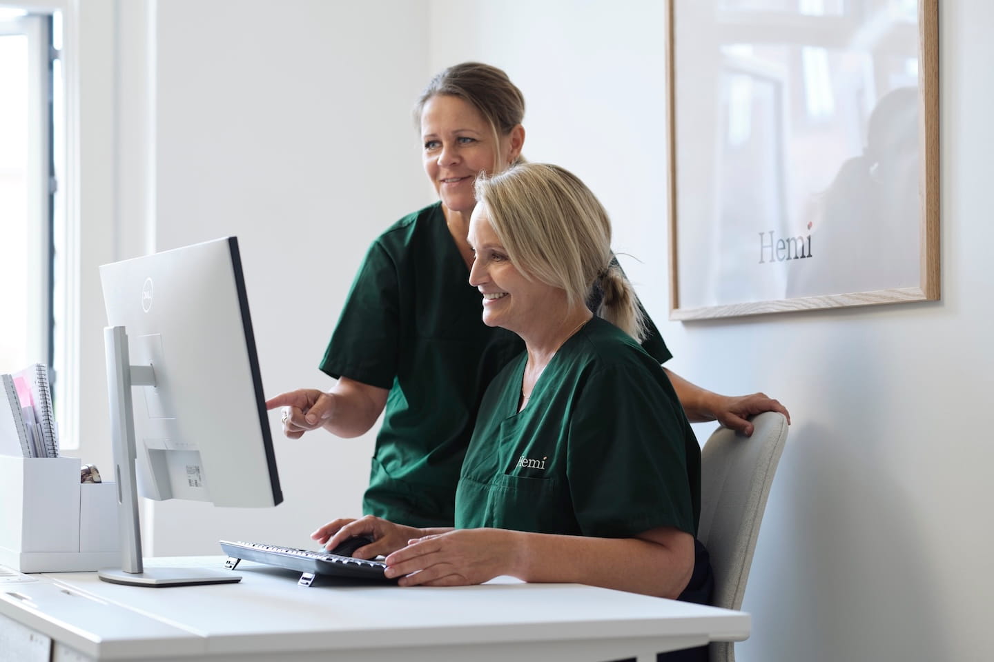 Twee vrouwen in groene medische kleding werken samen aan een computer in een lichte kantoorruimte.