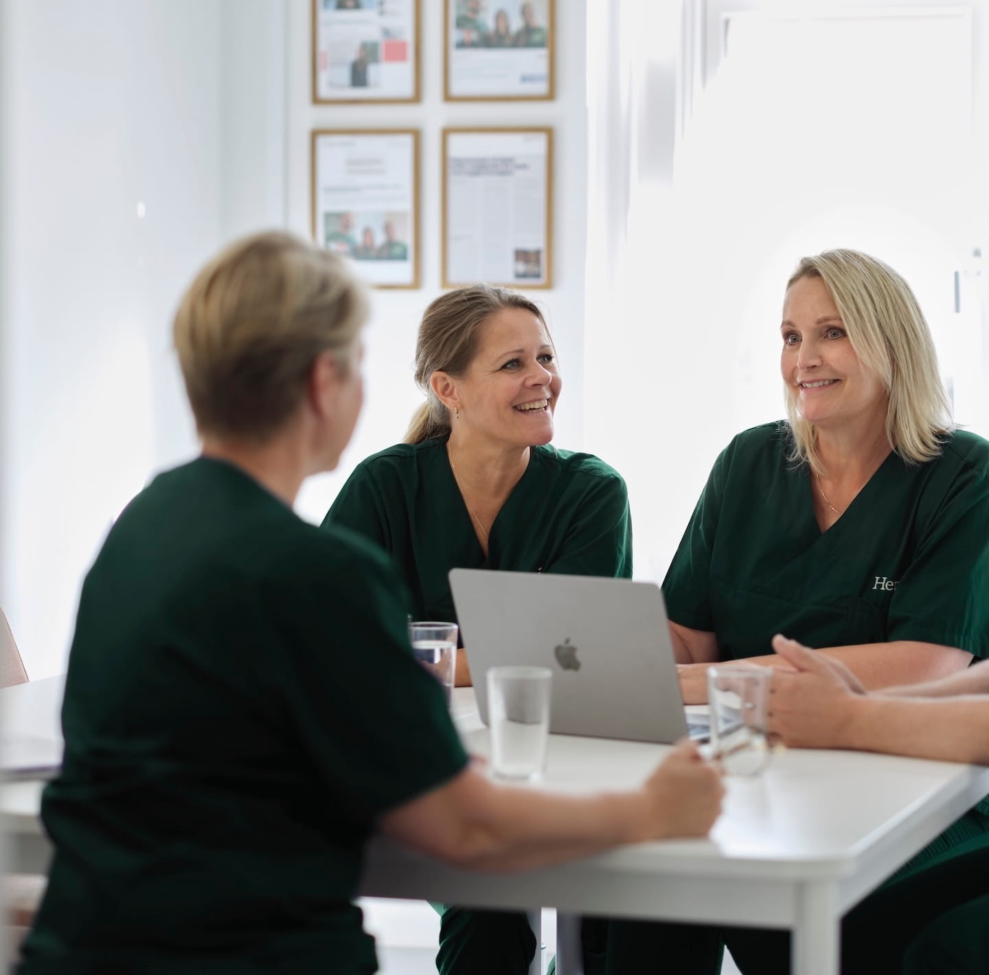 Drie vrouwen in groene medische kleding hebben een vergadering rond een witte tafel met een laptop en glazen water in een lichte kamer.