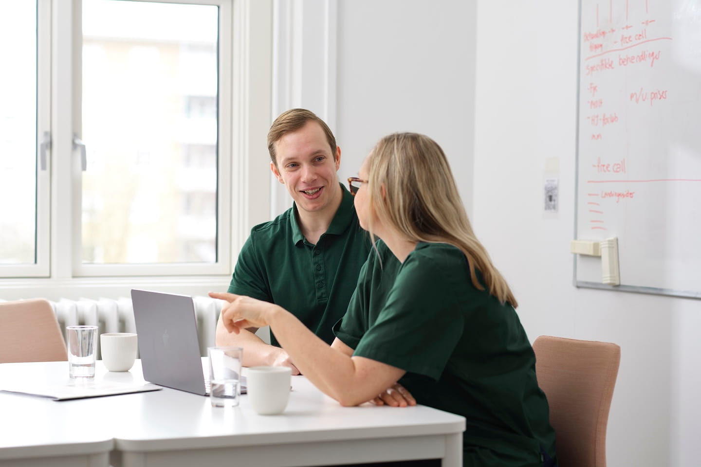 Twee medische professionals in groene kleding praten aan een bureau met een laptop en koffiekopjes.