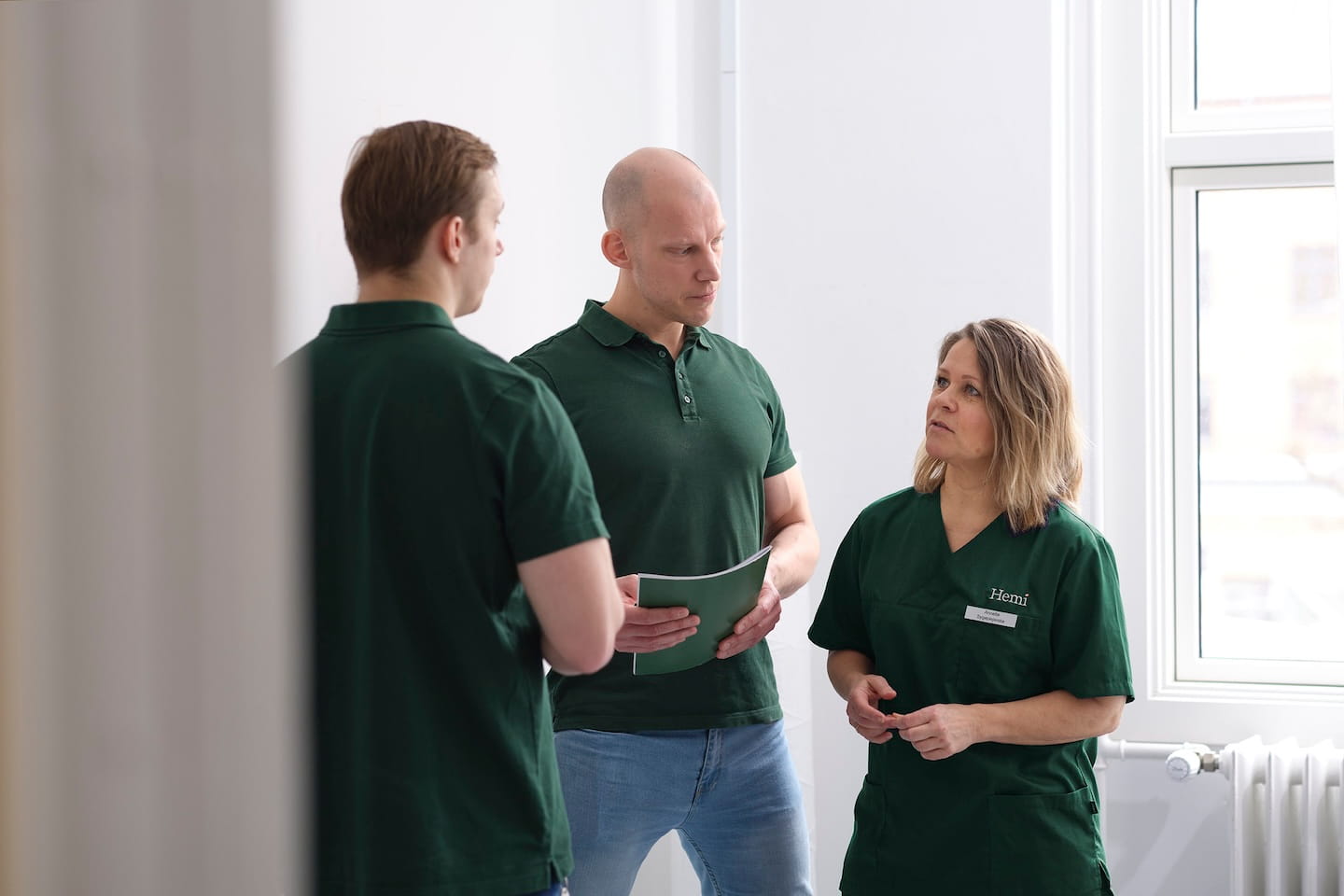 Drie mensen in groene kleding in gesprek in een lichte kamer bij een raam.