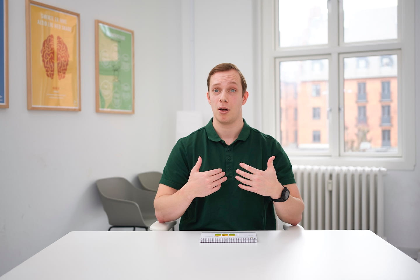 Man in groen poloshirt die zit aan tafel in een lichte kamer met posters aan de muur en spreekt terwijl hij zijn handen op zijn borst houdt.