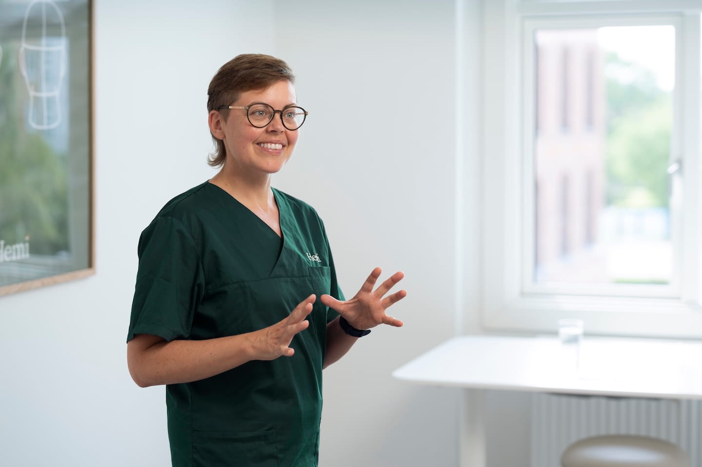 Vrouw in groene medische kleding met bril staat en praat in een lichte, moderne kamer met een raam en een tafel.