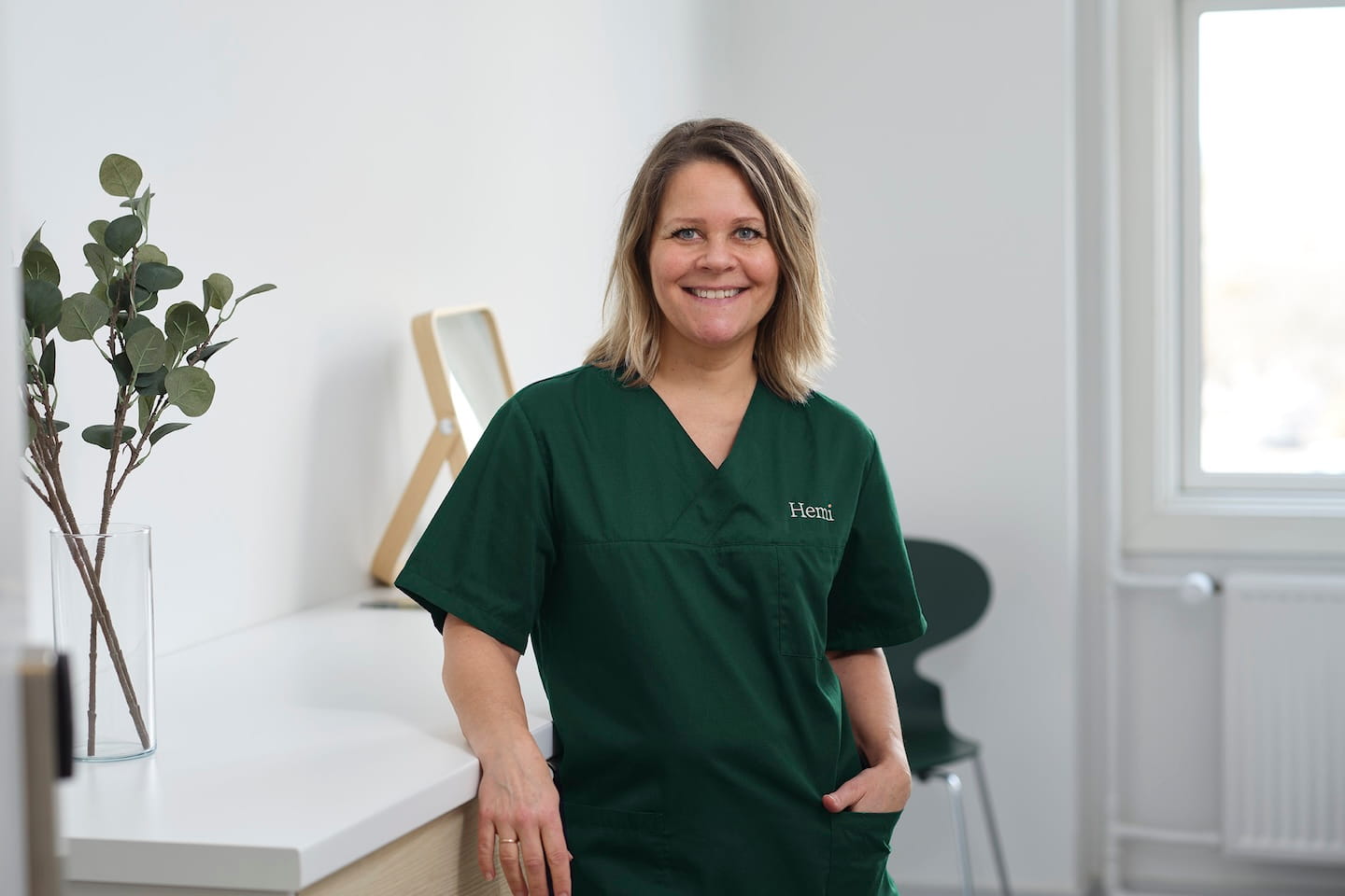 Vrouw in groene medische scrubs met het logo 'Hemi', lachend in een lichte kamer met een raam en een plant op de achtergrond.