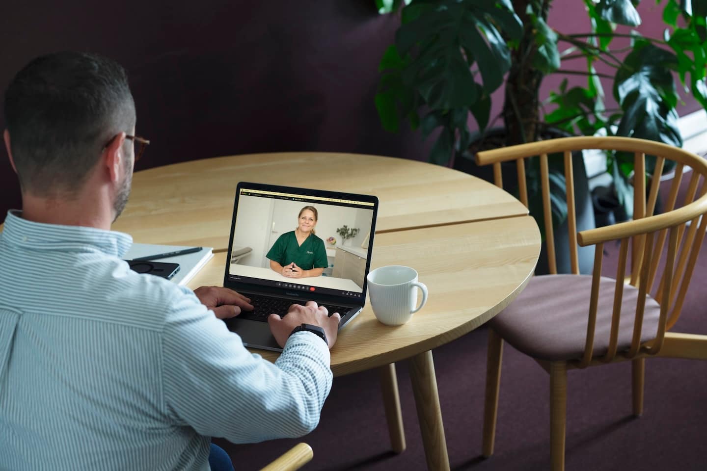 Man met bril zit aan een houten tafel en voert een videoconsult met een vrouw in groene medische kleding op zijn laptop.