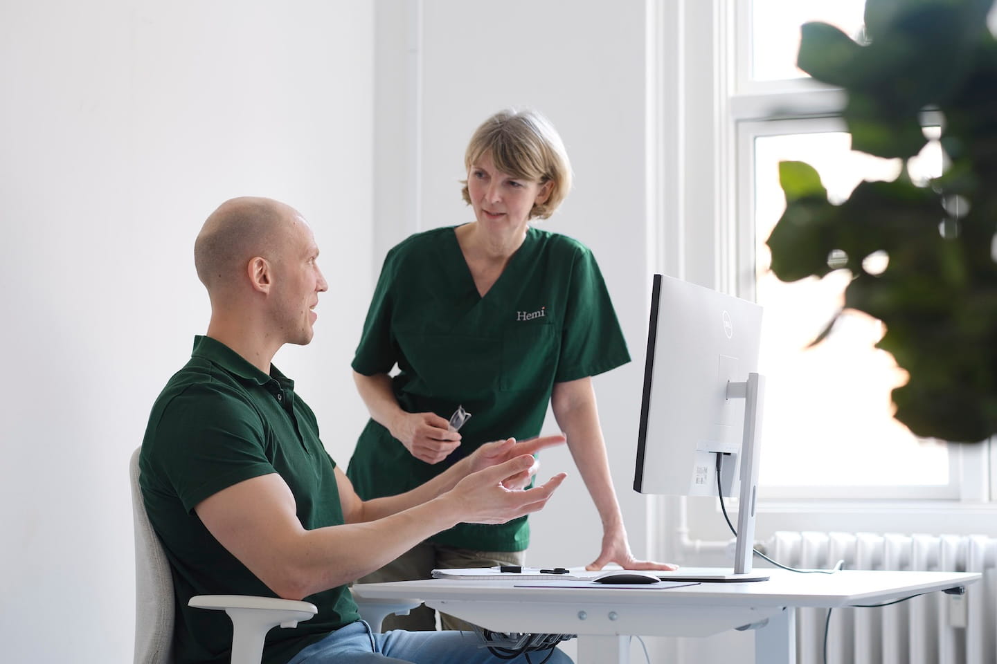 Twee mensen bespreken iets bij een computerscherm in een lichte kantoorruimte, de vrouw draagt een groen uniform met de naam Hemi.