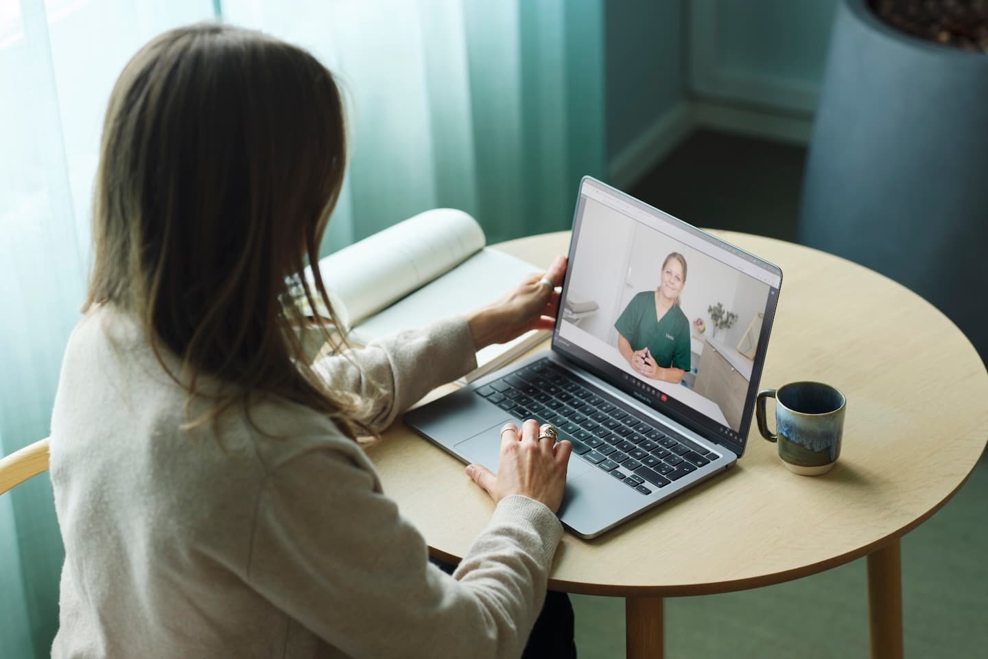Vrouw zit aan een houten tafel met laptop en heeft een videocall met een medisch professional in groene kleding.